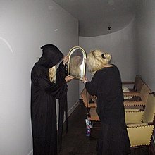 Dylan Brady and Laura Les wear black robes and hold a mirror.