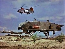 A colossal four-legged walking vehicle with two clawed arms is overflown by a twin-rotor military helicopter.