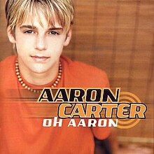 A male teenager with blonde hair is wearing an orange t-shirt and a bead necklace. The artist's name is colored in black and orange, and the album title is colored in white.