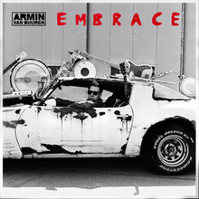 Armin Van Buuren (seen wearing sunglasses and his head facing towards the viewer) driving a rustic muscle car with musical instruments stacked on top of it