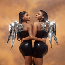 Chloe and Halle with shiny, metallic wings on their backs, both wearing latex dresses, standing and holding each other by one arm in front of a dusty background