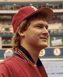A smiling young man with feathered blond hair wearing a red baseball jacket and red baseball cap
