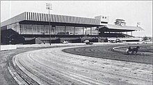 Picture of Rosecroft's track and old grandstand. A horse is racing on the track. To the right, there are several rows of bleachers with a metal tent above for protection.