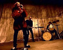 A female singer and a male bassist and drummer perform against a golden backdrop.