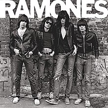 Four men standing against a graffiti-covered wall. Each man has a black leather coat, blue jeans, and brown hair. At the top of the black-and-white image, "RAMONES" is spelled out in all caps.