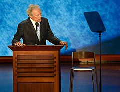 Man in coat and tie stands at a podium, looking towards a chair