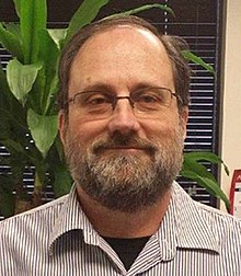 A balding brown-haired man with a beard, mustache and glasses facing the camera as he smiles slightly, wearing a narrow striped short with its collar open. Behind him is a window with closed blinds and a potted plant.