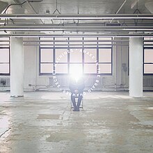 A man seated on a metal folding chair in an industrial building; his head is replaced by a bright white light