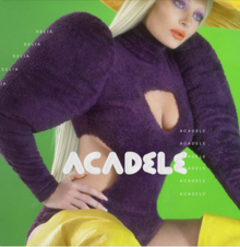 A shot of Delia in front of a green background, wearing a purple sweater along with yellow boots and a hat. The song's title is superimposed on her in white capital letters.