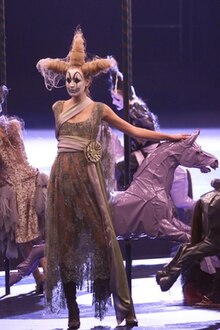 A woman wearing dark clown makeup, a wig shaped into three points, and a silver dress poses standing on a carousel