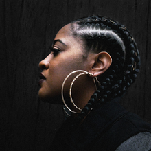 High contrast image of left side of Rapsody's face in profile with large cornrows and two large hoop earrings against a dark background
