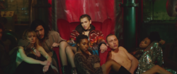 Dua Lipa sitting on a red chair in a warhouse, surrounded by models sitting on the floor.