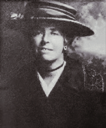 Cunnington in semi-formal dress as a young woman, wearing a wide-brimmed hat, a cardigan and a white blouse