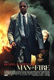 A man in a suit and sunglasses is walking away from a fiery blaze, his arm is held out to guard a small blonde girl