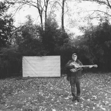 The album cover is a black-and-white photograph of a cricket field with a sight screen in the distance, off-center to the left. Off-center to the right and in the distance Fred Frith is standing playing a guitar. There is no text on the cover.