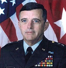 Color photo of a man in formal military clothing in front of an American flag