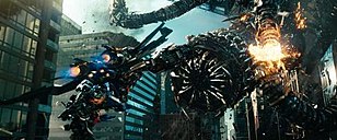 In a city full of skyscrapers, a robot wearing jet thrusters on his back holding a cannon flies toward a large snake-like robot. A tentacle of the larger robot is exploding.