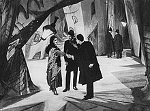 A black-and-white film still. Three figures stand facing each other on a heavily stylized street set.